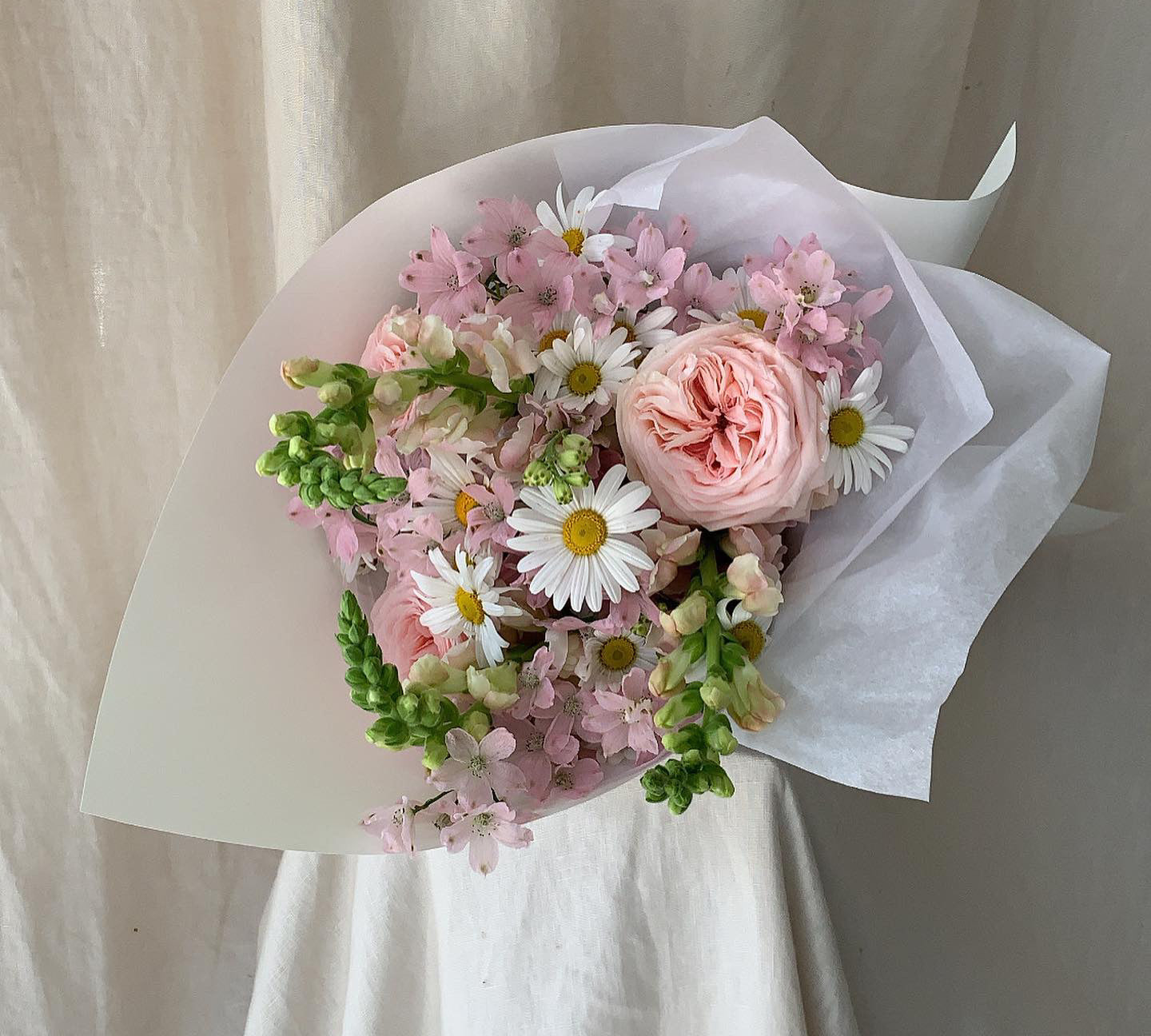 Sweet posie of Australian grown, seasonal blooms including daisies, pale pink fragrant roses, pale pink mini delphinium wrapped in stone coloured paper sitting on linen covered plinth. 