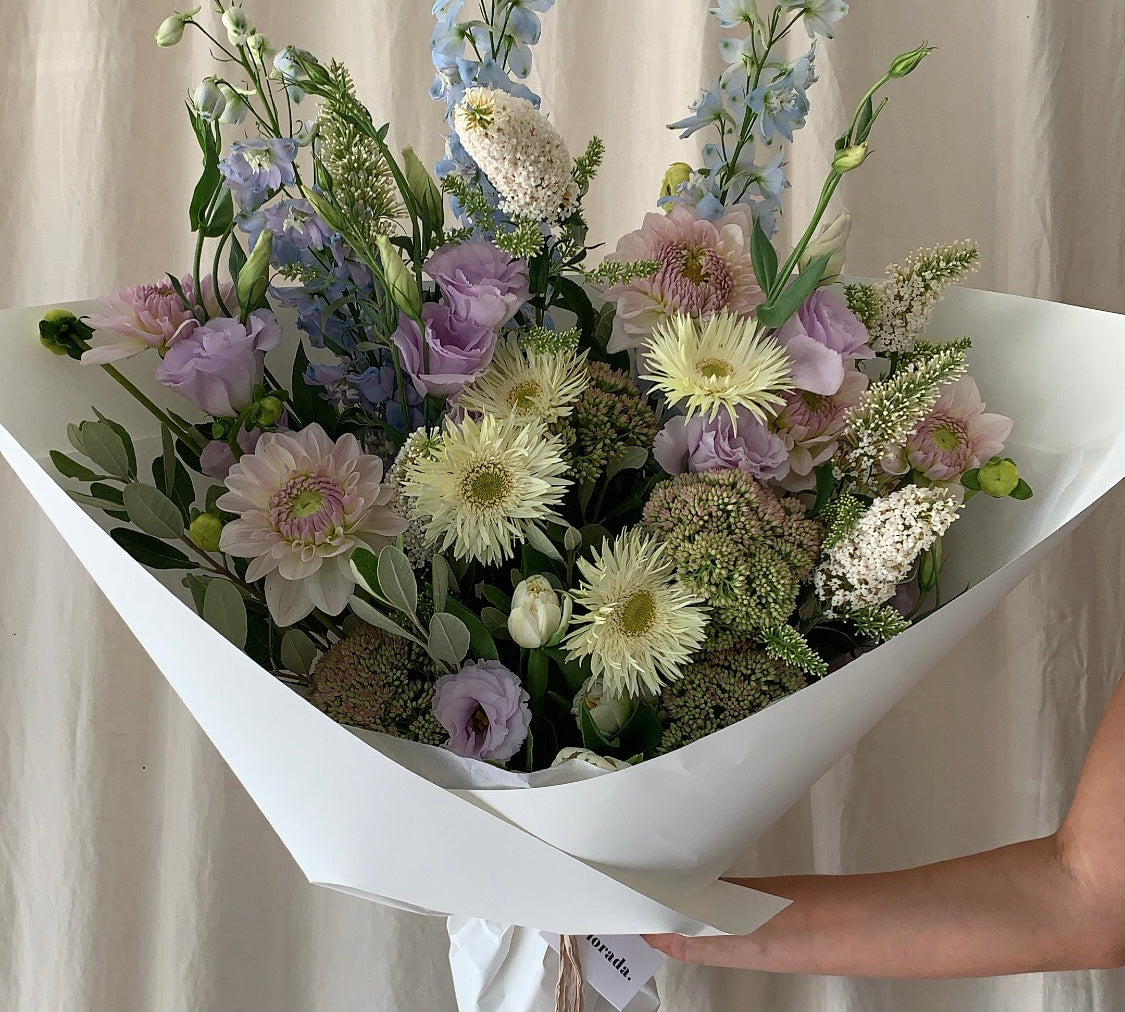 An arrangement of soft, pretty, garden-style blooms including dahlia, gerbera, lisianthus, delphinium and fragrant buddleia, wrapped in Florada low-waste paper and silk ribbon sourced from Australian growers.