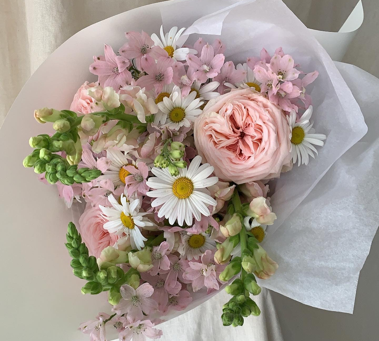 Close up of posie including daisies, pale pink fragrant roses, pale pink mini delphinium wrapped in stone coloured paper sitting on linen covered plinth. 
