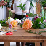 Workshop table with flowers, foliage, scissors scattered as people make flower arrangements.