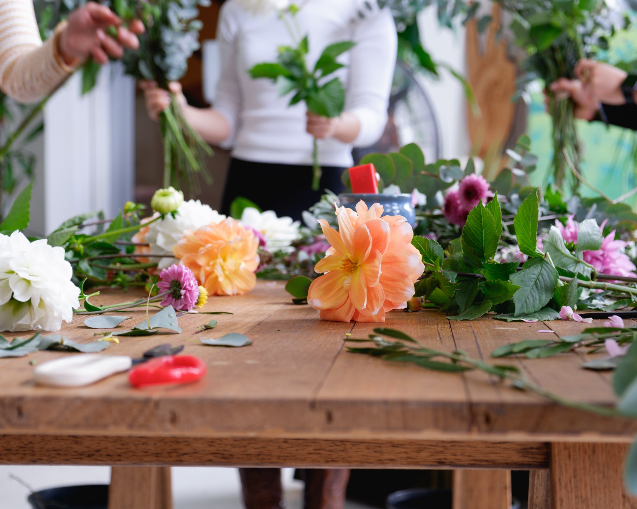 Workshop table with flowers, foliage, scissors scattered as people make flower arrangements.