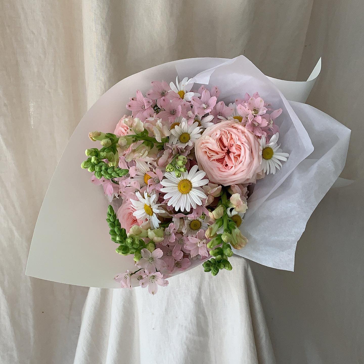 Sweet posie of Australian grown, seasonal blooms including daisies, pale pink fragrant roses, pale pink mini delphinium wrapped in stone coloured paper sitting on linen covered plinth. 